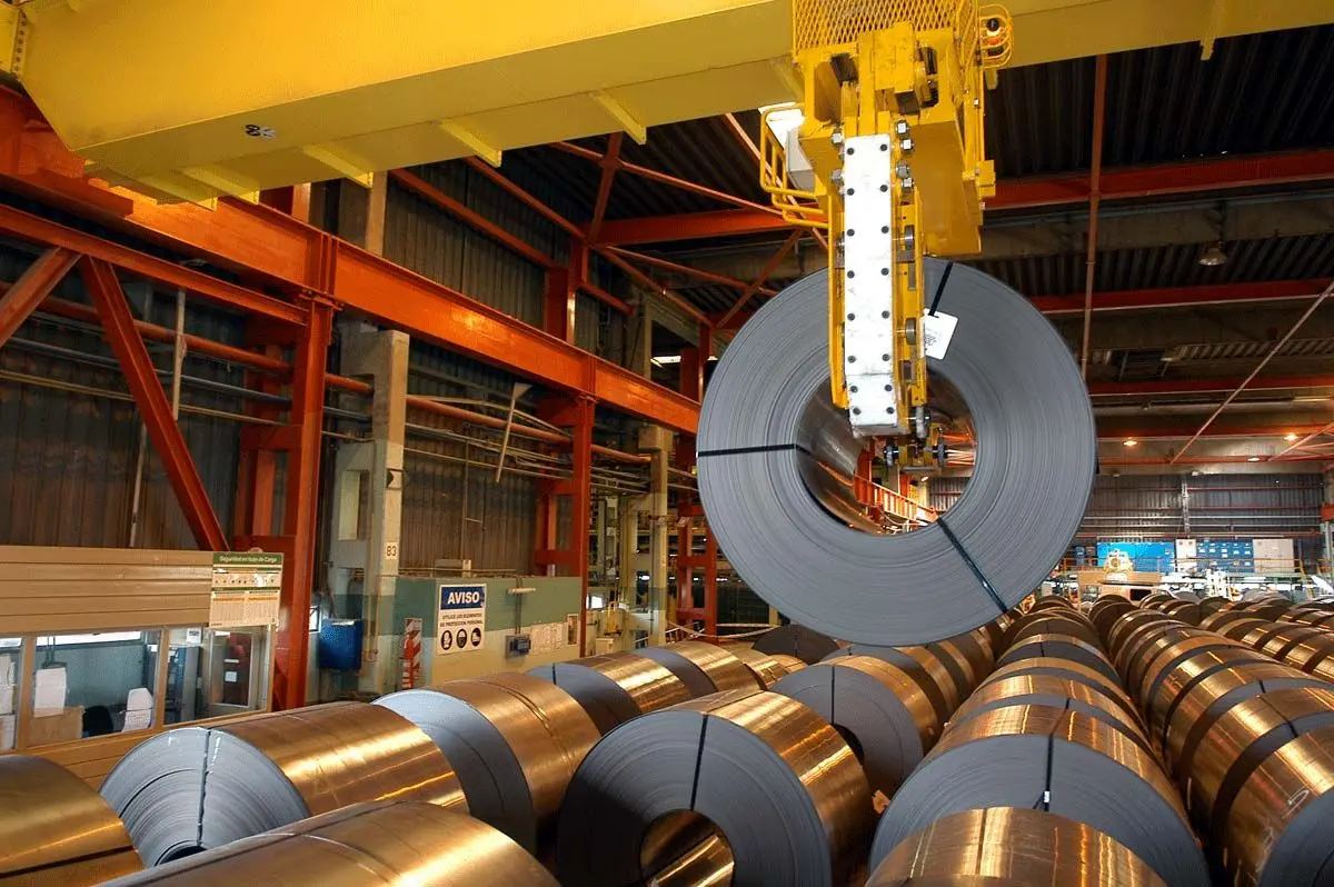 Overhead crane lifting a steel coil in a metal processing mill, with rows of coiled metal.